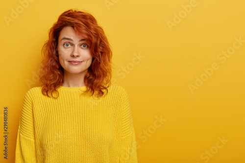 Foto Portrait of young ginger woman has natural beauty, dressed in yellow knitted jumper, dimples at cheeks looks at camera with eyebrows raised, likes bright clothes