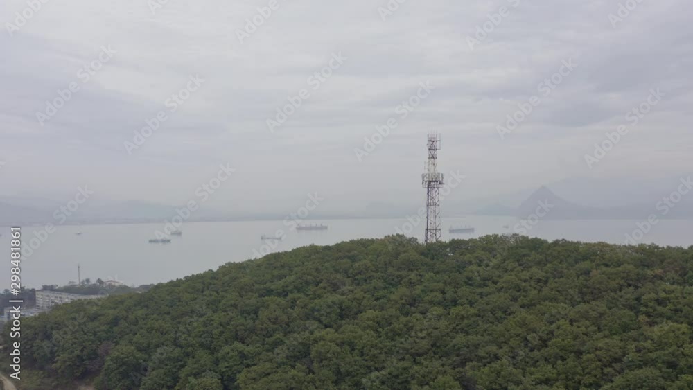 Radio Telecom tower on top of a green forest mountain with the reveal of bay area with ships at anchorage and mountain ridge in far distance. Aerial shot Radio Telecom tower on top of a green forest mountain with the reveal of bay area with ships at anchorage and mountain ridge in far distance. Aerial shot