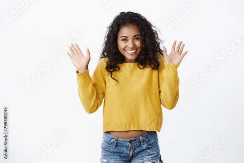 Happy pretty african american girl with curly hair showing excitement and happiness, greeting friends, waving raised palms hello, hi gesture, finally can wear new yellow sweater autumn chilly day