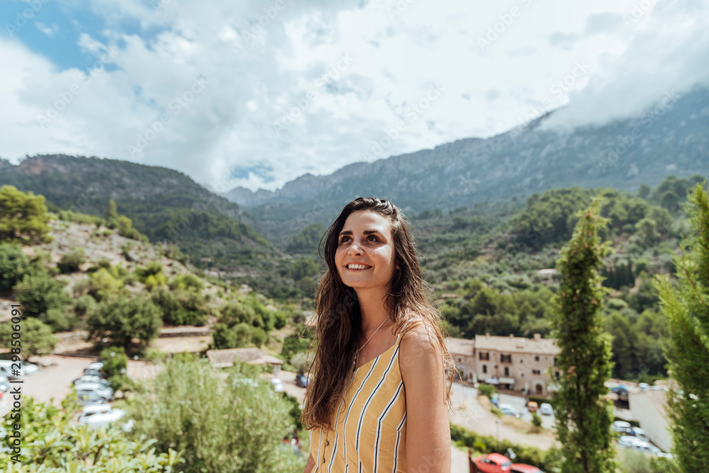 Beautiful Caucasian girl dressed casual, smiles and looks up at the sky, with bushy plants and trees in the background landscape, in the picturesque village of Fornalutx, Mallorca, Spain. Vertical