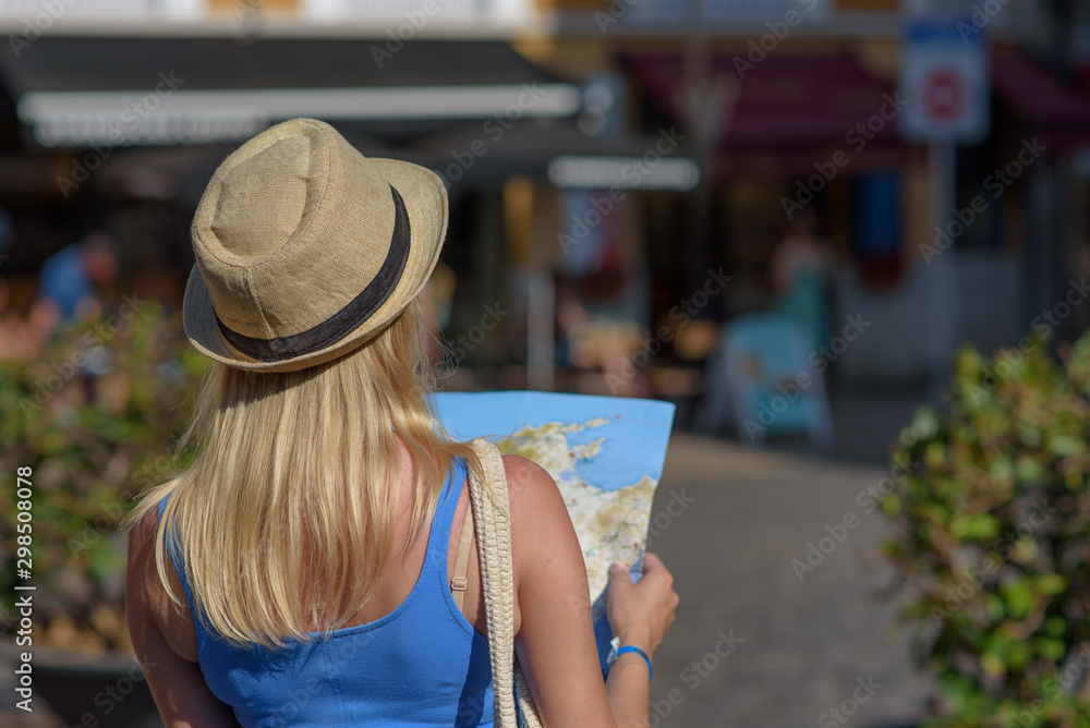 Fototapeta premium Young blonde beautiful girl in sun hat with map in her hands walking along European city, trying to find right way. Back view.