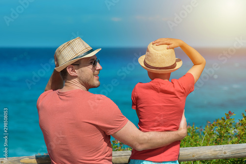 Dad and son in sun hats standing on cozy nature balcony with the sea view. They happily spend summer vacations abroad travelling and enjoying different views.