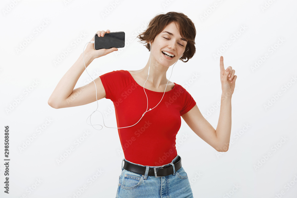 Girl lip sync having amusing carefree time carried away music bits dancing raise index fingers close eyes singing along favorite song hold smartphone wear earbuds standing white background Girl lip sync having amusing carefree time carried away music bits dancing raise index fingers close eyes singing along favorite song hold smartphone wear earbuds standing white background