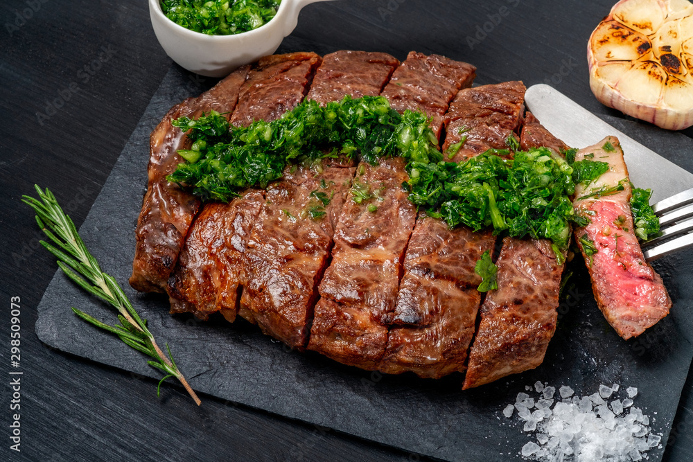 Ready to eat black Angus beef rib eye steak sliced with herbs, garlic and sauce on slate Board. Ready meal for dinner on black wooden background