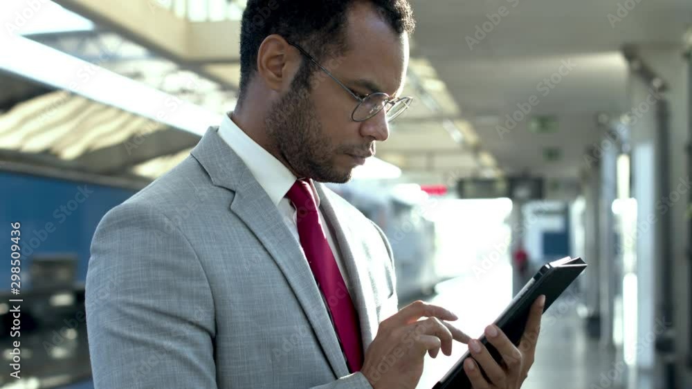 Businessman with digital tablet at train station. Focused young African American businessman in formal wear and eyeglasses standing and using tablet pc at railway station. Digital device concept Businessman with digital tablet at train station. Focused young African American businessman in formal wear and eyeglasses standing and using tablet pc at railway station. Digital device concept