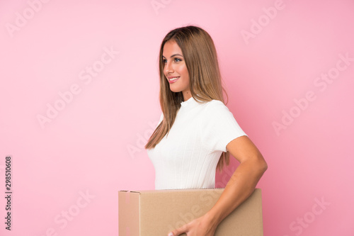 Young blonde woman over isolated pink background holding a box to move it to another site in lateral position © luismolinero