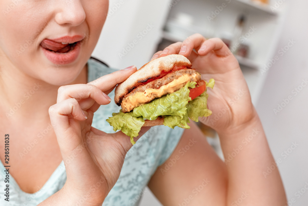 Junk Food. Chubby girl sitting at kitchen eating hamburger smiling playful close-up side view