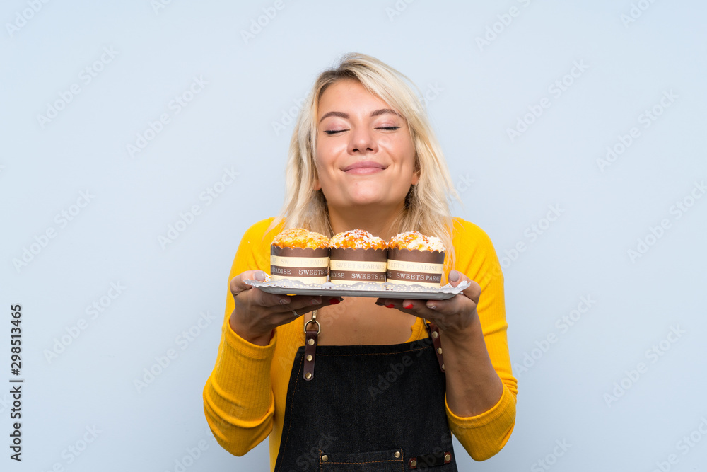 Young blonde woman over isolated background holding mini cakes enjoying the smell of them Young blonde woman over isolated background holding mini cakes enjoying the smell of them