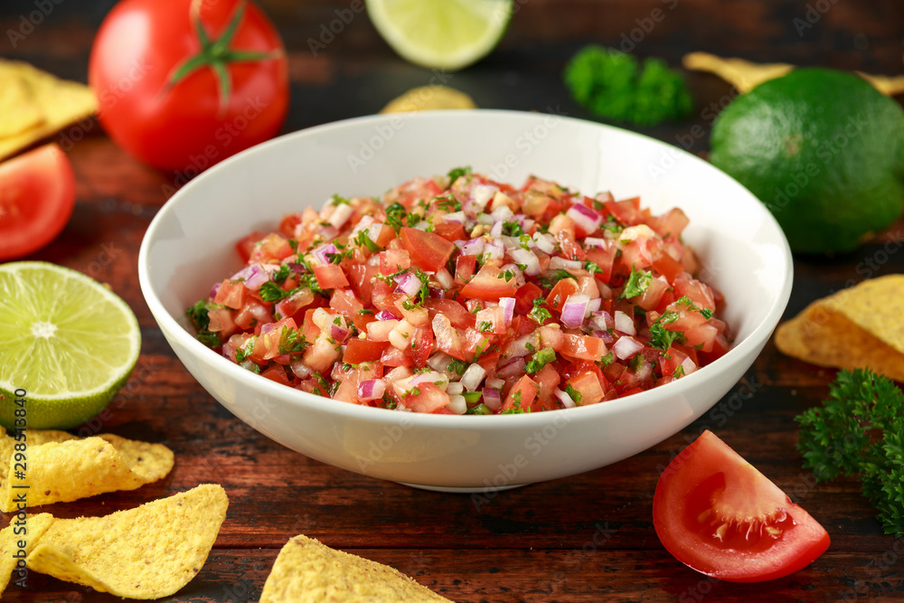Mexican Tomato Salsa in white bowl with lime, red onion, jalapeno pepper, parsley and tortilla chips on wooden table Mexican Tomato Salsa in white bowl with lime, red onion, jalapeno pepper, parsley and tortilla chips on wooden table