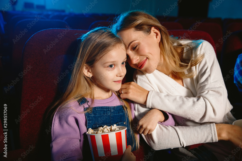 Naklejka premium Caucasian mother and daughter watching a film at a movie theater, house or cinema. Looks expressive, astonished and emotional. Sitting alone and having fun. Relation, love, family, childhood, weekend.