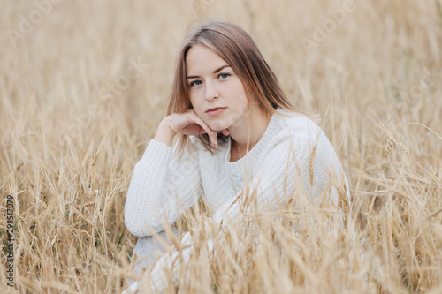 Beautiful young girl in a white sweater sits in a wheat field and looks thoughtfully at the camera.