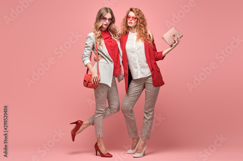 Two fashionable woman sisters in Trendy autumn red elegant outfit, stylish hair, makeup hugging. Gorgeous friends in jacket, heels on pink. Sensual model girl, stylish pastel fashion accessories