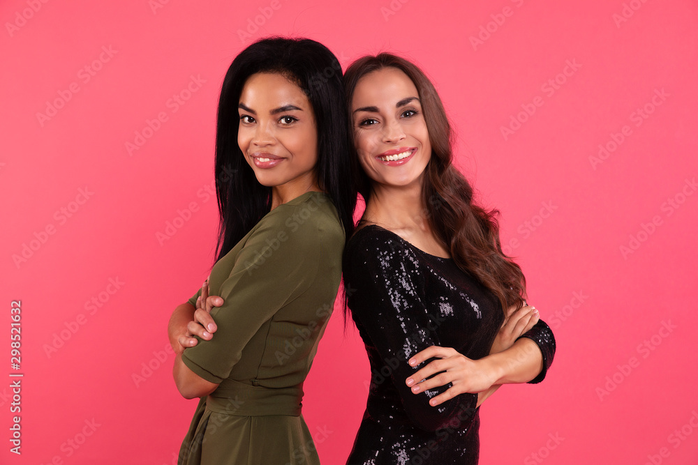 Party for everybody. Two girls in party dresses are standing back to back with their arms folded, looking at the camera with dazzling smiles and sparking eyes. Party for everybody. Two girls in party dresses are standing back to back with their arms folded, looking at the camera with dazzling smiles and sparking eyes.