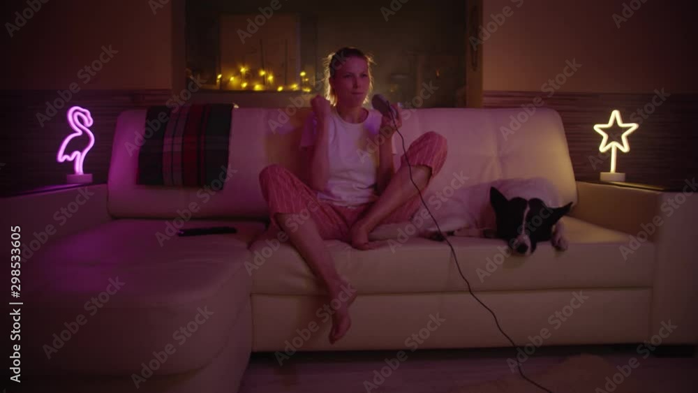 Beautiful Excited Young Gamer Girl with Dog Sitting on a Couch, Playing and Winning in Video Games on a Console. She Plays with a Wireless Controller. Cozy Room is Lit with Warm and Neon Light. Beautiful Excited Young Gamer Girl with Dog Sitting on a Couch, Playing and Winning in Video Games on a Console. She Plays with a Wireless Controller. Cozy Room is Lit with Warm and Neon Light.