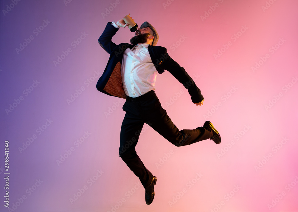 Full length portrait of happy jumping man wearing office clothes in neon light isolated on gradient background. Emotions, ad concept. Hurrying up, late for work or sale, shopping. Drinks coffee to go.