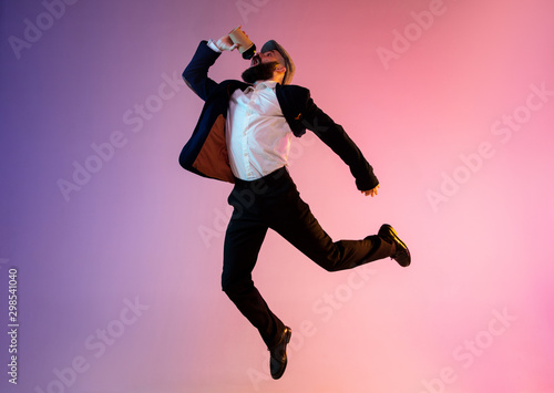 Full length portrait of happy jumping man wearing office clothes in neon light isolated on gradient background. Emotions, ad concept. Hurrying up, late for work or sale, shopping. Drinks coffee to go.
