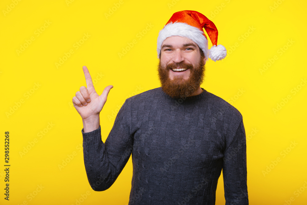 Image of handsome man with beard pointing away at copyspace and wearing santa claus hat Image of handsome man with beard pointing away at copyspace and wearing santa claus hat