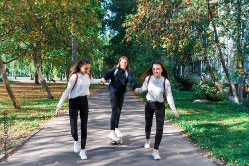 Three girls schoolgirls teenagers 13-15 years old, autumn summer day city, ride skateboard, happy smiling laughing, casual clothes, rest after school break. Emotions joy fun and enjoyment.