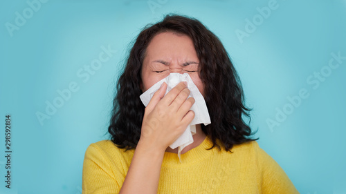 Burnette woman blowing nose into tissue. Flu cold or allergy symptom. Sick woman girl sneezing in tissue on blue. Health care. photo