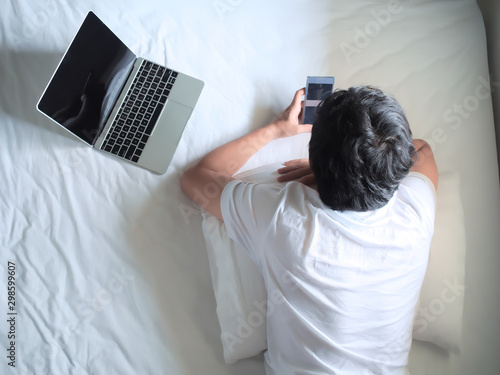Top view of young man using phone and laptop on the bed in bedroom.