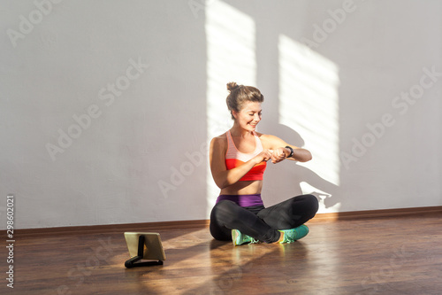Satisfied sporty woman with bun hairstyle and in tight sportswear looking at smartwatch, checking time or pulse after workouts, tablet on floor. indoor studio shot illuminated by sunlight from window