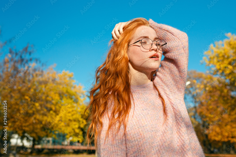 Portraits of a charming red-haired girl with a cute face. Girl posing in autumn park in a sweater and a coral-colored skirt. The girl has a wonderful mood
