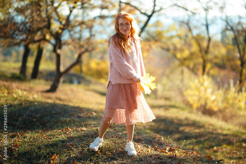 Portraits of a charming red-haired girl with a cute face. Girl posing in autumn park in a sweater and a coral-colored skirt. In the hands of a girl a yellow leaf Portraits of a charming red-haired girl with a cute face. Girl posing in autumn park in a sweater and a coral-colored skirt. In the hands of a girl a yellow leaf