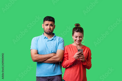 Portrait of funny young man in casual wear standing with crossed arms, looking resentful at camera, upset about woman using phone and ignoring him. isolated on green background, indoor studio shot
