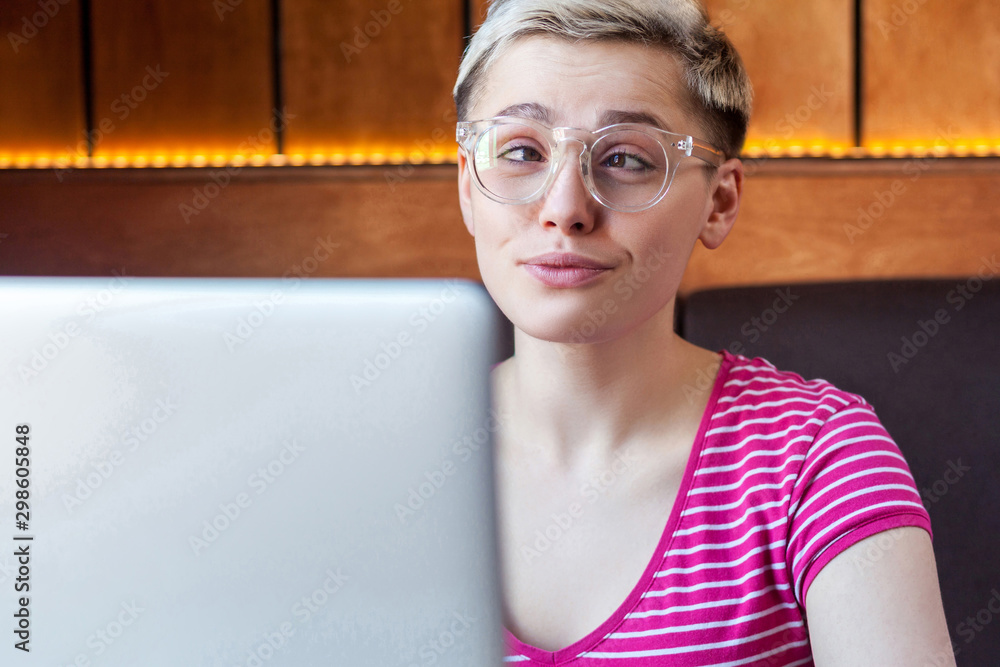 Closeup portrait of happy beautiful childish young girl with short hair in pink t-shirt and eyeglasses is sitting in cafe and working on laptop, crossed eyed like stupid, looking at camera, indoor Closeup portrait of happy beautiful childish young girl with short hair in pink t-shirt and eyeglasses is sitting in cafe and working on laptop, crossed eyed like stupid, looking at camera, indoor