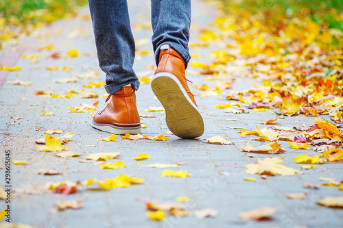 Back view on the feet of a man in brown boots walking along the sidewalk strewn with fallen leaves. The concept of turnover of the seasons of the year. Weather background