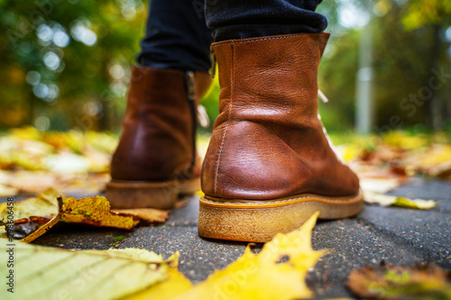 legs of a woman in black pants and brown boots walking in a park along the sidewalk strewn with fallen leaves. The concept of turnover seasons. Weather background