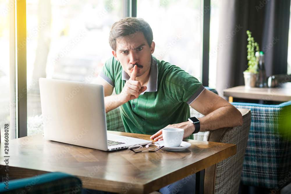 Shhh, Be quiet! Young angry businessman in green t-shirt sitting and working on laptop, looking at camera with finger on lips and command to be silent. indoor shot near big window at daytime. Shhh, Be quiet! Young angry businessman in green t-shirt sitting and working on laptop, looking at camera with finger on lips and command to be silent. indoor shot near big window at daytime.