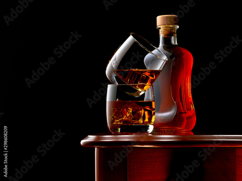 Two glasses of whiskey and ice on the wooden table. The glasses are stacked on top of each other. In the background, a closed bottle of whiskey. Low key.