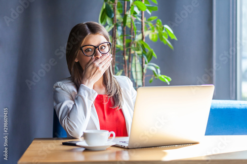 Canvas Print I will be quiet! Portrait of shocked beautiful brunette young woman in glasses sitting, looking at camera with big eyes and closing her mouth with hand