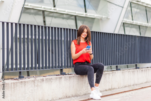 Mobile internet at public places. Portrait of positive charming brunette woman in stylish red shirt sitting near metal fence  texting on phone or scrolling social media  wireless network. outdoor shot
