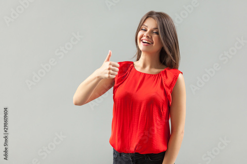 Like! Portrait of satisfied beautiful brunette young woman in red shirt standing, thumbs up and looking at camera with toothy smile and joyful face. indoor, studio shot, isolated on gray background.
