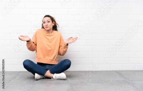 Young woman with curly hair sitting on the floor having doubts with confuse face expression