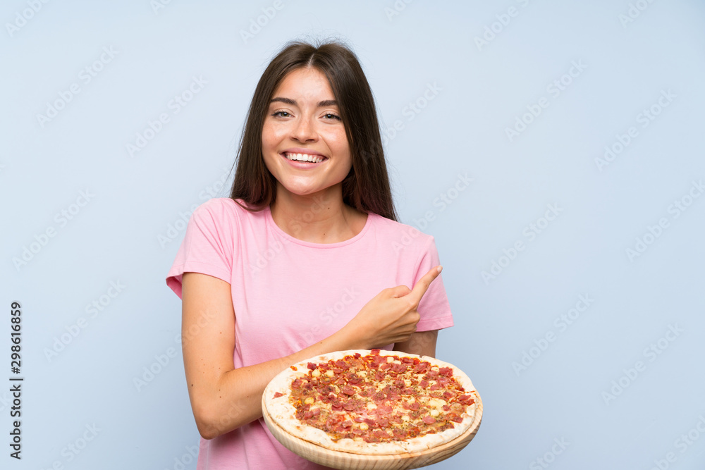 Pretty young girl holding a pizza over isolated blue wall pointing to the side to present a product