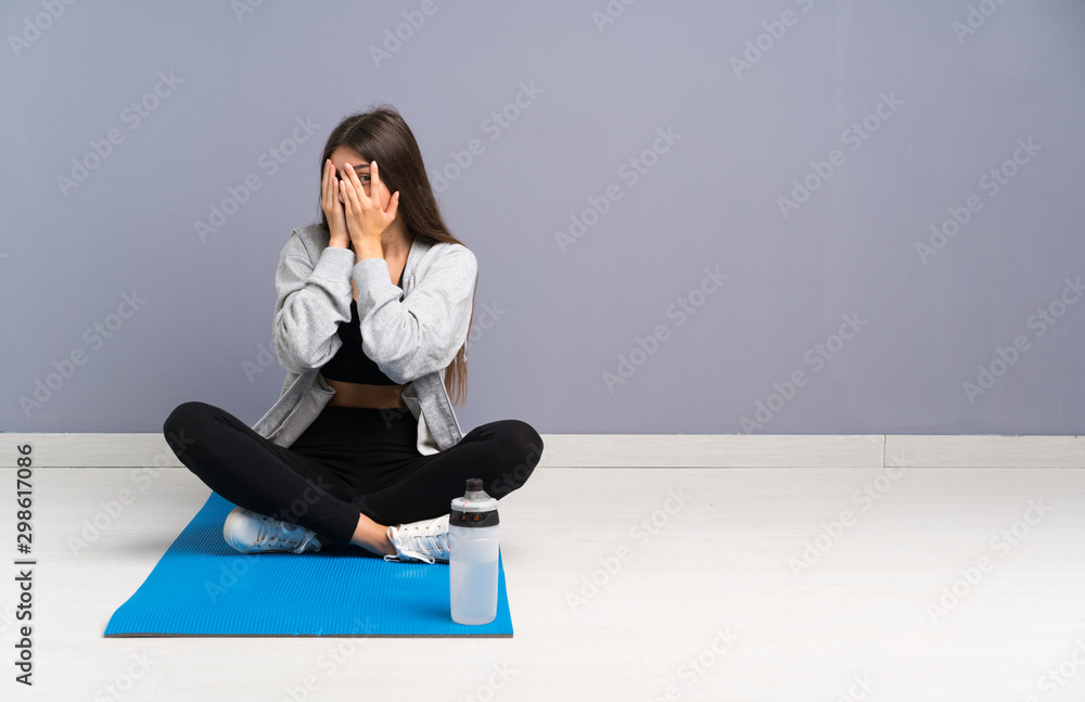 Young sport woman sitting on the floor with mat covering eyes and looking through fingers Young sport woman sitting on the floor with mat covering eyes and looking through fingers