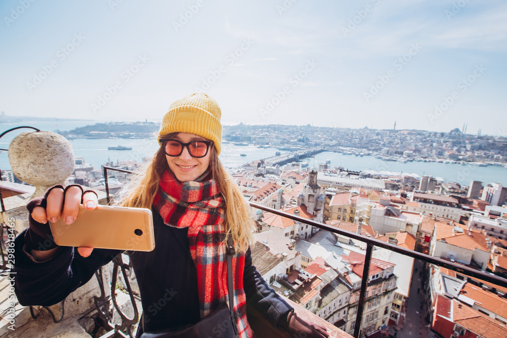 Young smiling girl on the background of Istanbul panorama on a sunny spring day. A girl in yellow hats takes a selfie on the phone in Istanbul from the observation deck of the Galata Tower.