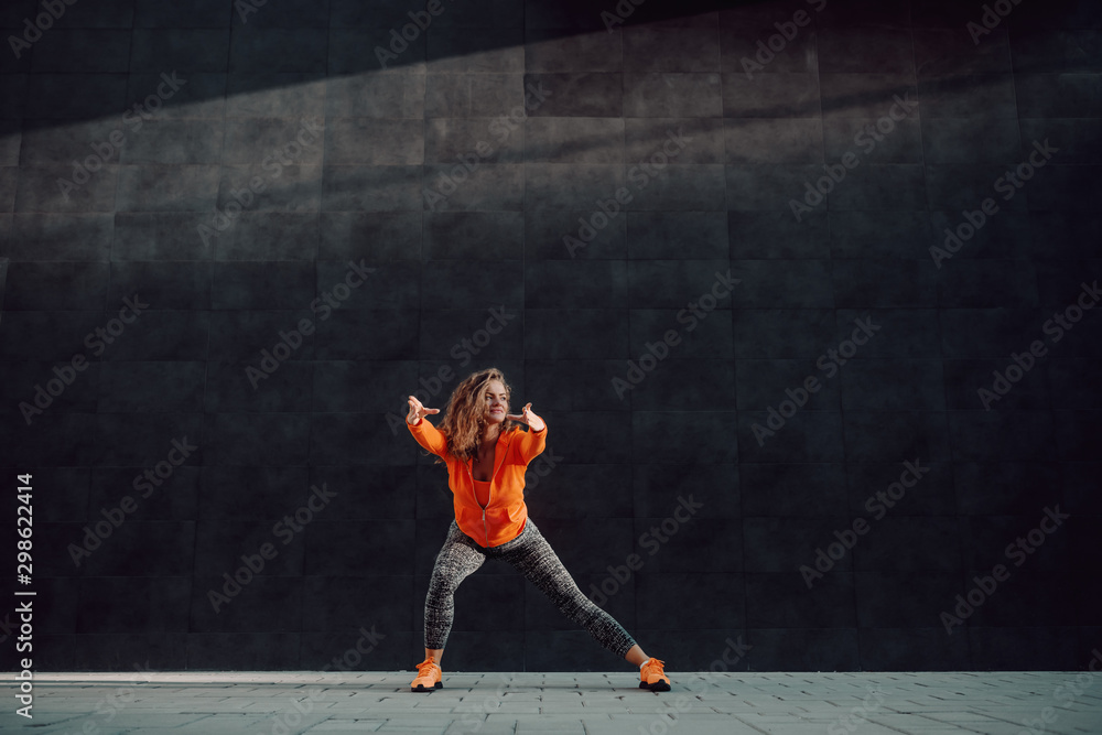 Attractive caucasian sporty woman in sportswear and with curly hair doing warm up exercises for legs while standing in front of black wall.