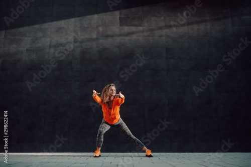 Attractive caucasian sporty woman in sportswear and with curly hair doing warm up exercises for legs while standing in front of black wall.