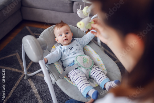 Adorable baby boy lying in bouncer an looking at his mother. Mother entertaining her loving son. Living room interior. photo