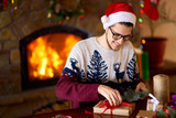 Happy man in a Santa hat creates gifts. The man made them himself and is going to give for the holidays. Concept of gifts and greetings for Christmas and New Year. Beautiful background with fireplace.