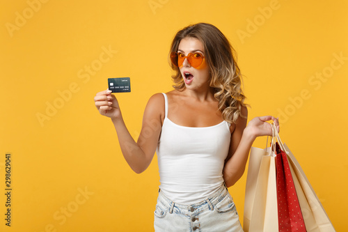 Shocked young woman girl in eyeglasses posing isolated on yellow orange background. People lifestyle concept. Mock up copy space. Holding package bag with purchases after shopping, credit bank card.