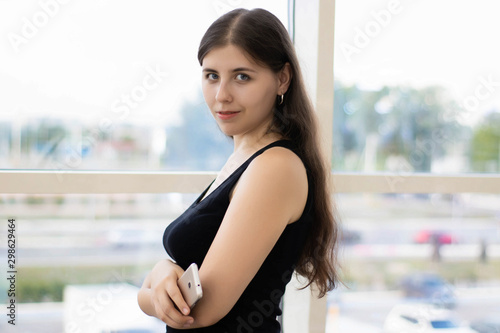 Free Download A young woman with long hair is standing against the window in the mall, holding a smartphone in her hands. - GFXLoad