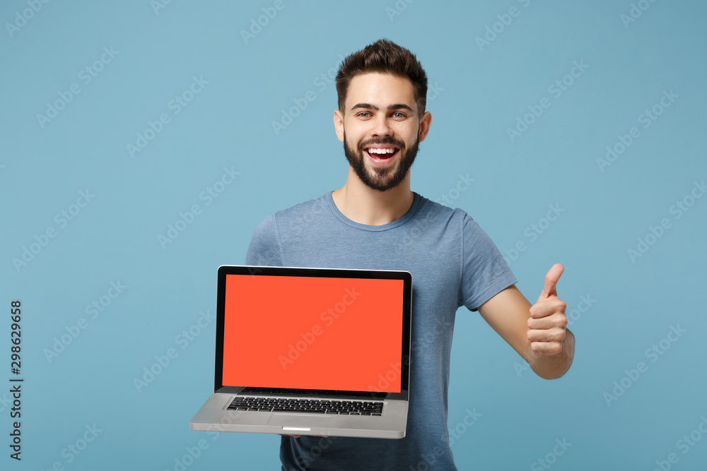 Young laughing man in casual clothes posing isolated on blue wall background in studio. People lifestyle concept. Mock up copy space. Hold laptop pc computer with blank empty screen, showing thumb up.