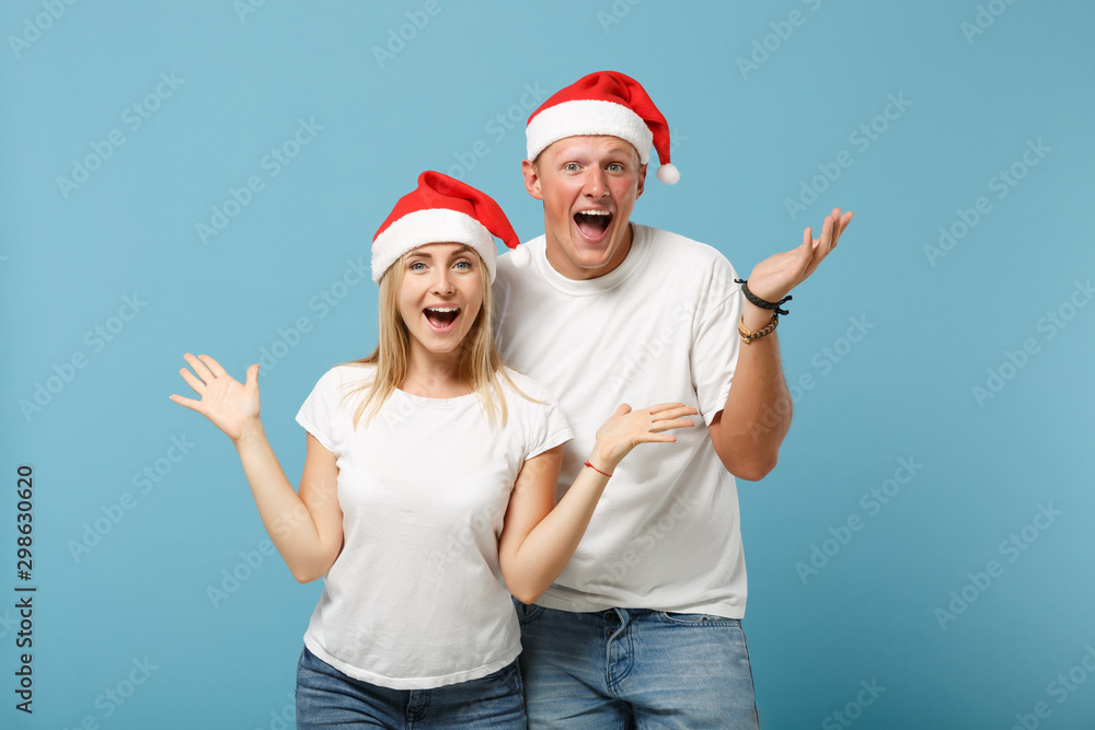 Obraz premium Excited young Santa couple friends guy girl in Christmas hat posing isolated on pastel blue background in studio. Happy New Year 2020 celebration holiday concept. Mock up copy space. Spreading hands.
