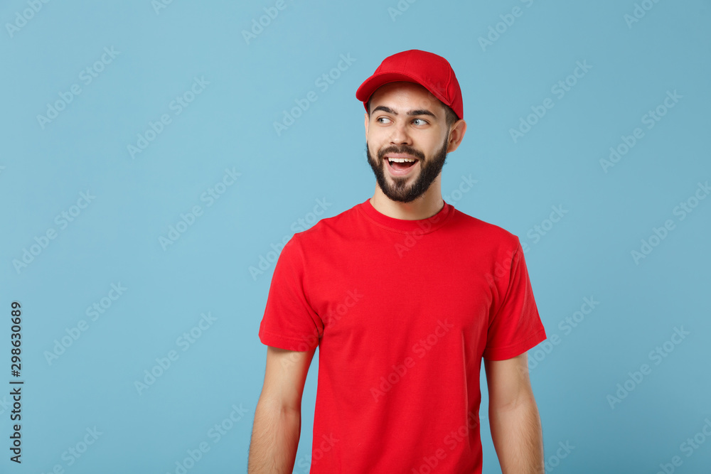 Delivery man in red uniform workwear isolated on blue wall background, studio portrait. Professional male employee in cap t-shirt print working as courier dealer. Service concept. Mock up copy space. Delivery man in red uniform workwear isolated on blue wall background, studio portrait. Professional male employee in cap t-shirt print working as courier dealer. Service concept. Mock up copy space.