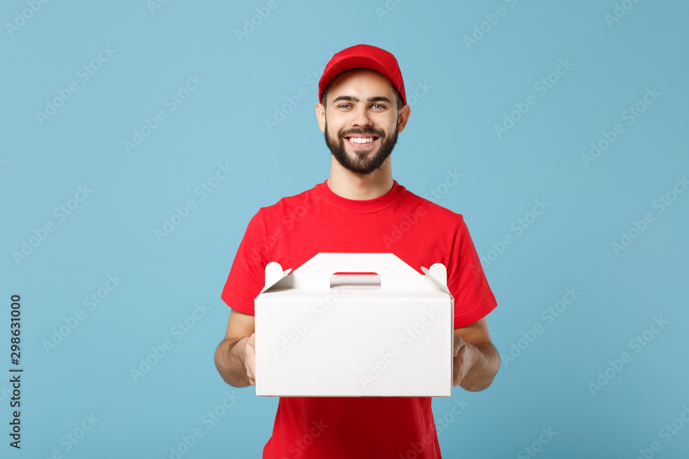 Delivery man in red uniform isolated on blue background, studio portrait. Male employee in cap t-shirt print courier hold cake dessert in unmarked cardboard box. Service concept. Mock up copy space. Delivery man in red uniform isolated on blue background, studio portrait. Male employee in cap t-shirt print courier hold cake dessert in unmarked cardboard box. Service concept. Mock up copy space.